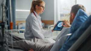 Image of a female neurologist speaking with a sick male patient resting in bed while using a tablet computer to show and explain brain scan results.