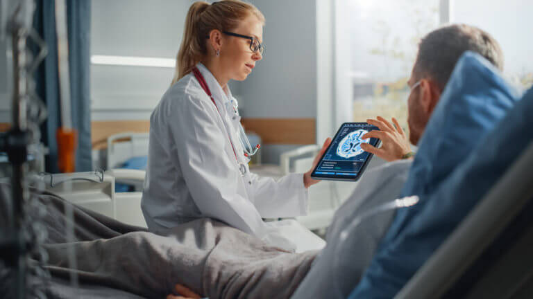 Image of a female neurologist speaking with a sick male patient resting in bed while using a tablet computer to show and explain brain scan results.