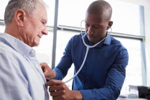 A male primary care doctor is listening to the chest of a senior male patient during a medical exam in the office.